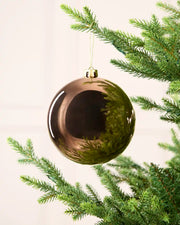 A large, glossy, metallic Mocha Christmas bauble ornament hanging from a green pine tree branch against a soft, off-white background.