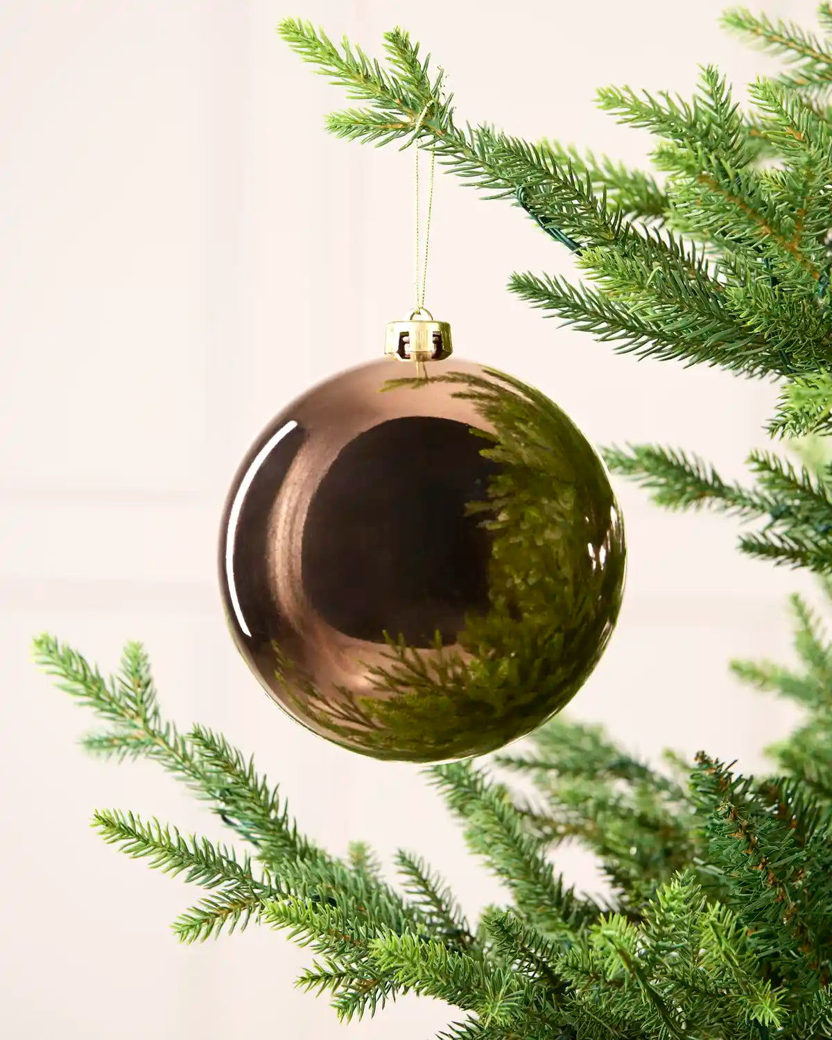 A large, glossy, metallic Mocha Christmas bauble ornament hanging from a green pine tree branch against a soft, off-white background.