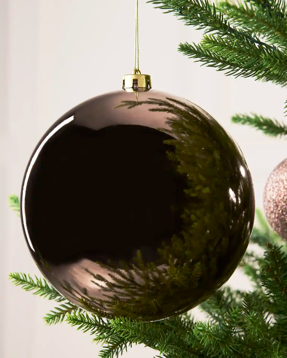 A large, glossy mocha Christmas bauble ornament hanging from a gold cap on a green Christmas tree branch, reflecting the surrounding pine needles and a soft light background.
