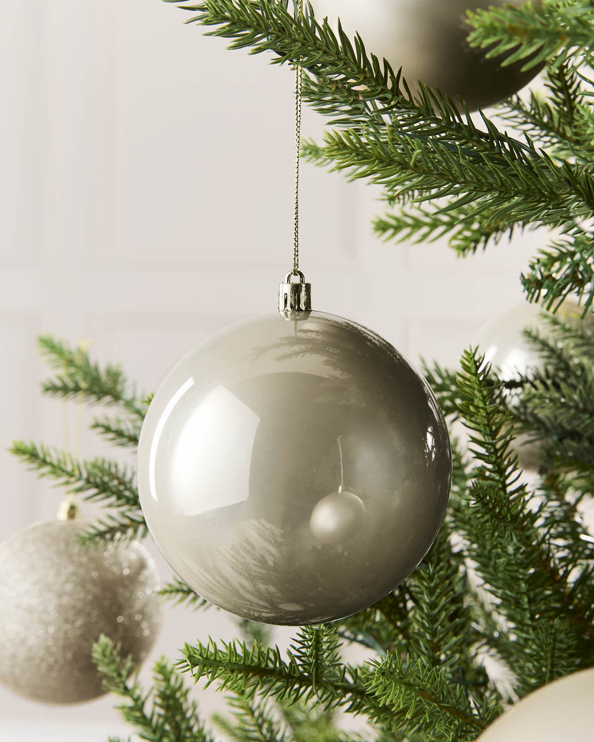 A large, glossy spherical Christmas ornament in a warm latte color hangs from a silver string on a green pine tree branch.