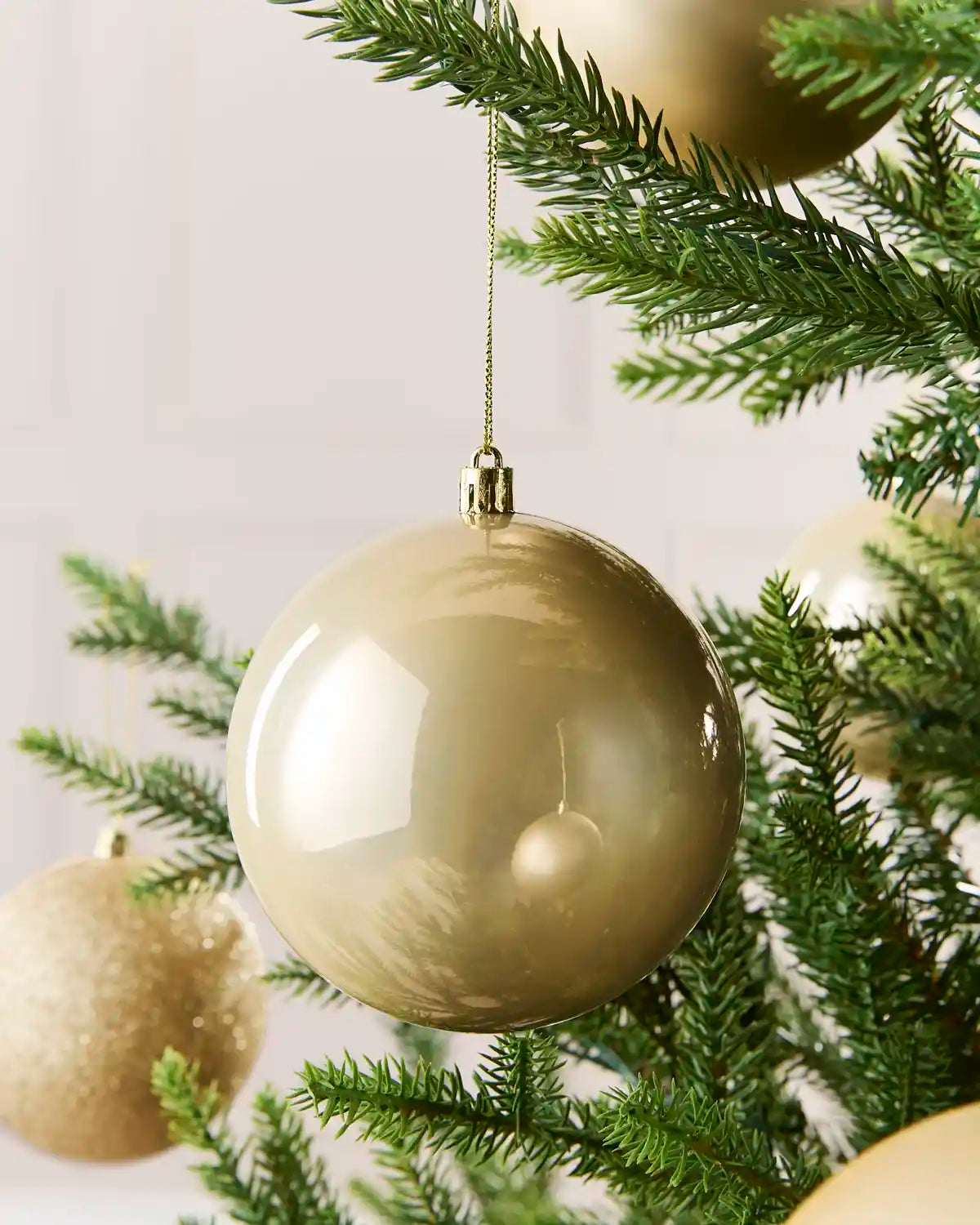 A large, glossy spherical Christmas ornament in a warm latte color hangs from a silver string on a green pine tree branch.
