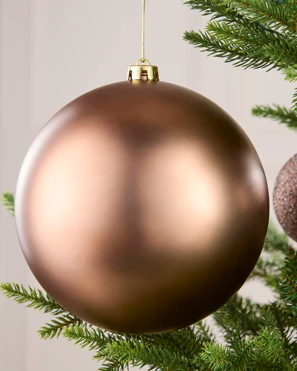 A large matte Mocha Christmas ball ornament hanging from a green pine tree branch against a soft light gray background.