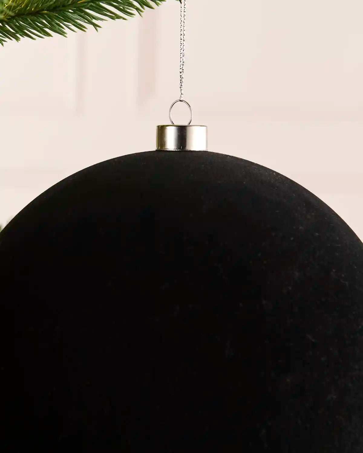A large matte black spherical Christmas ornament hanging from a silver cap with a thin metal chain, with pine branches visible at the top edge against a soft light background.