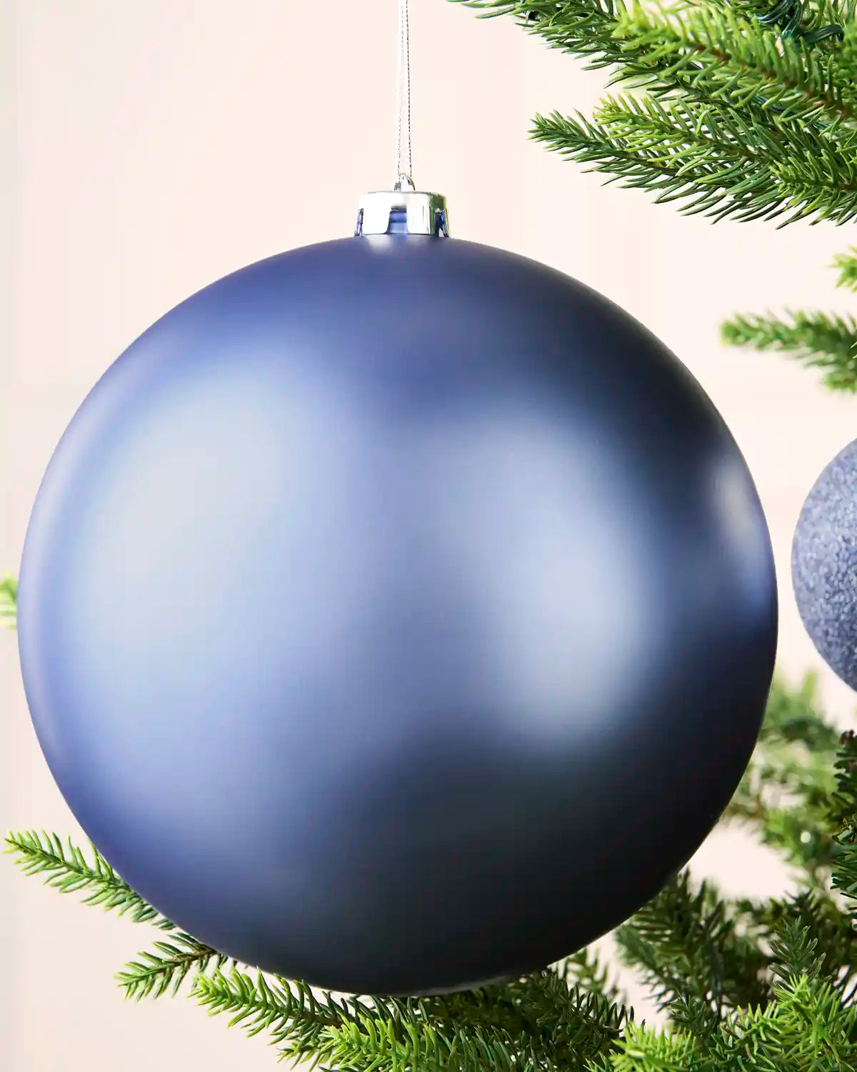 A large matte blue Christmas bauble hanging from a silver cap on a tree branch, with green pine needles and part of another ornament visible.