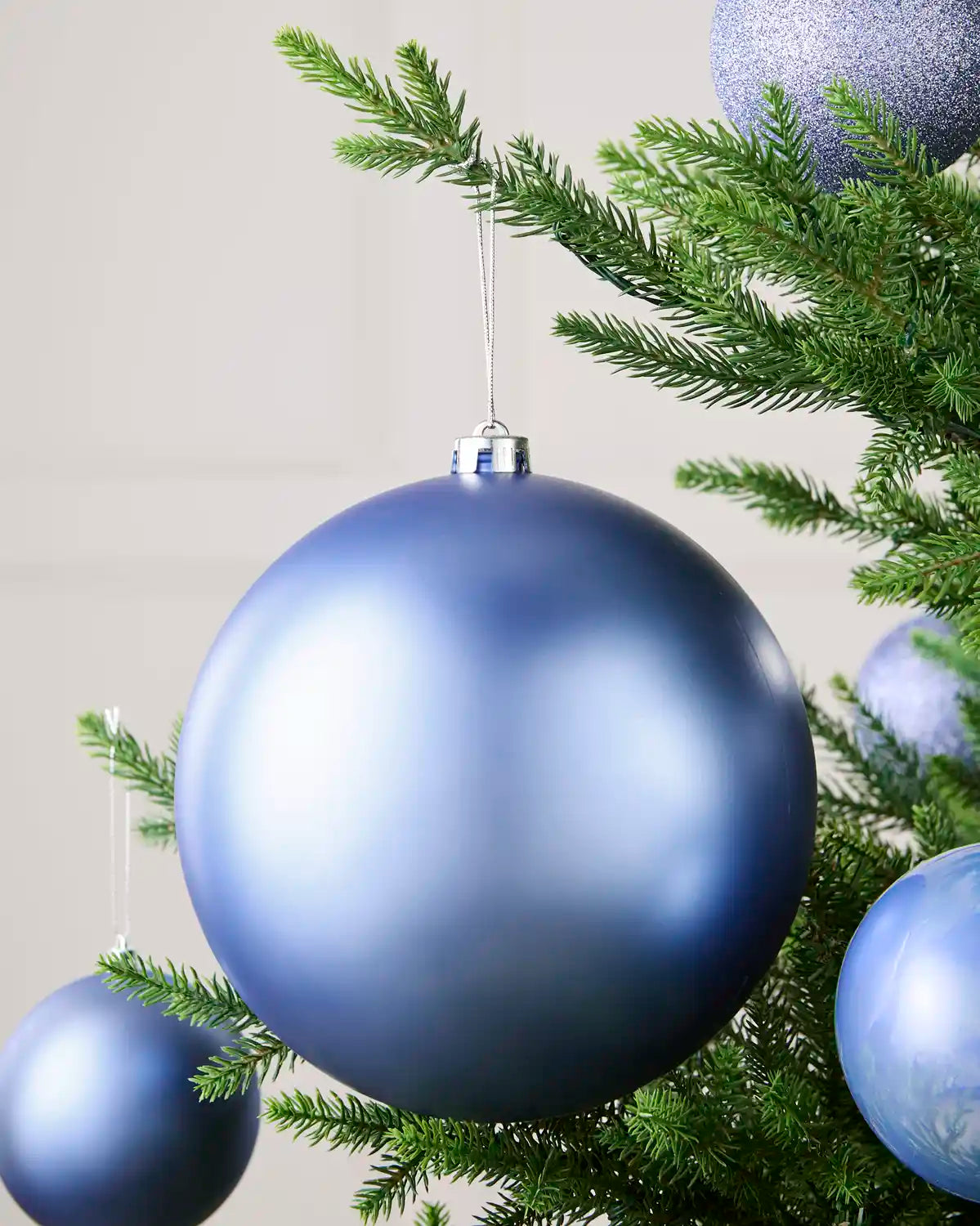 A large matte blue Christmas bauble ornament hanging from a green pine tree branch, with other matching blue and silver ornaments visible around it.