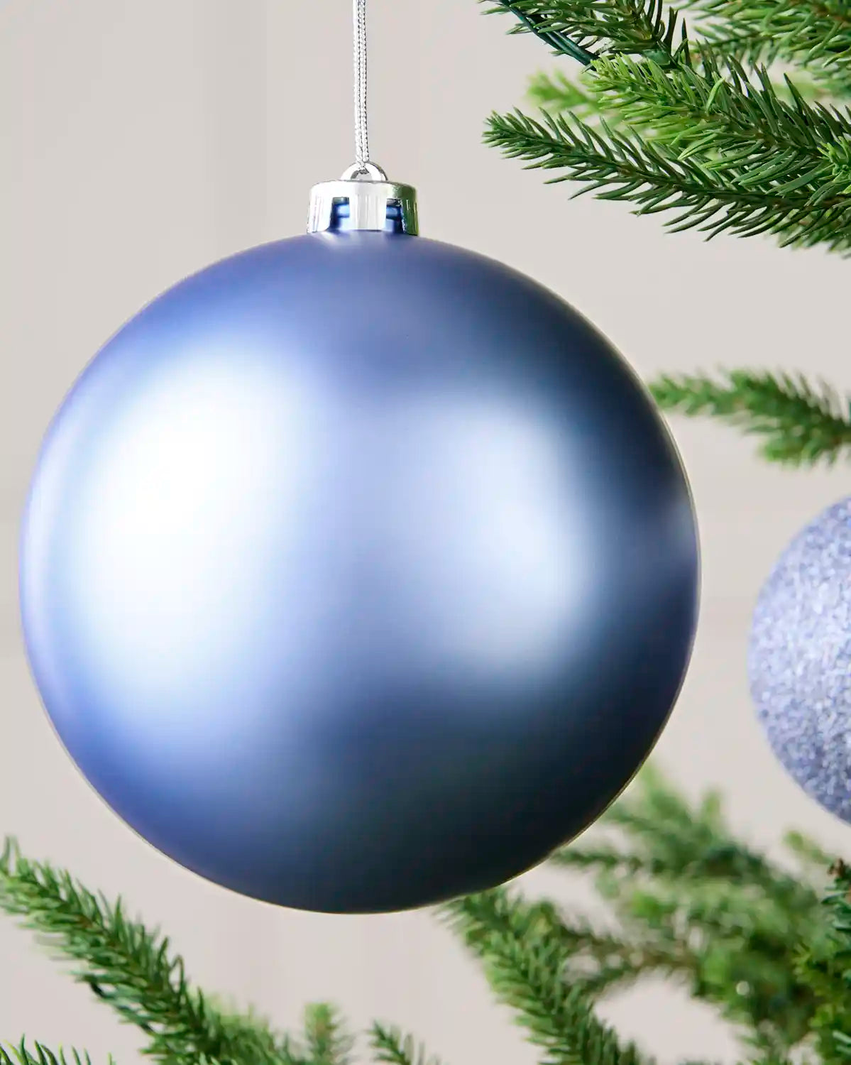 A large matte blue Christmas bauble ornament hanging from a silver cap on a tree branch, with green pine needles and part of another silver ornament visible in the background.