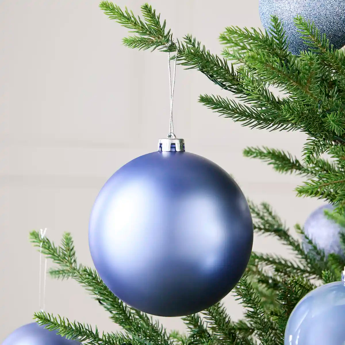 A large matte blue Christmas tree ornament hanging from a green pine branch, with other blue ornaments partially visible around it.