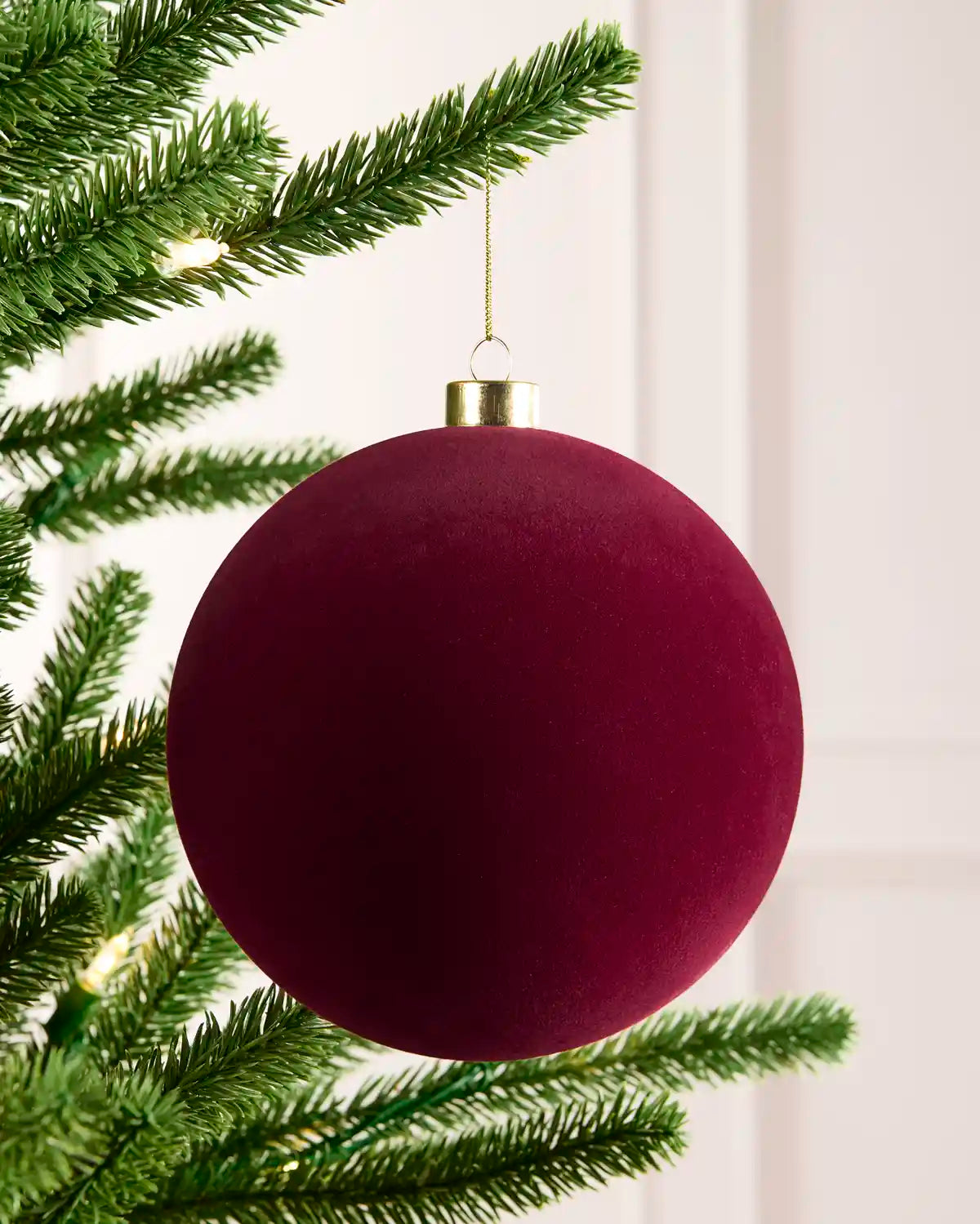 A large, matte burgundy Christmas tree ornament hanging from a green pine tree branch with warm white string lights in the background.