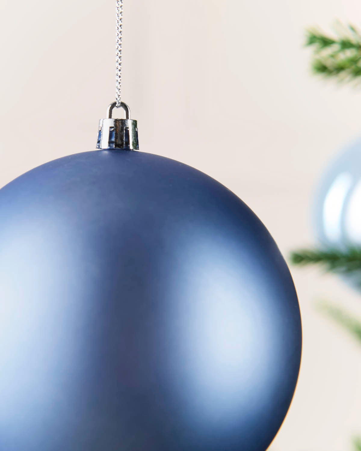 A large matte dark ice blue Christmas ornament hanging from a silver chain, shown in a close-up with a softly blurred background.