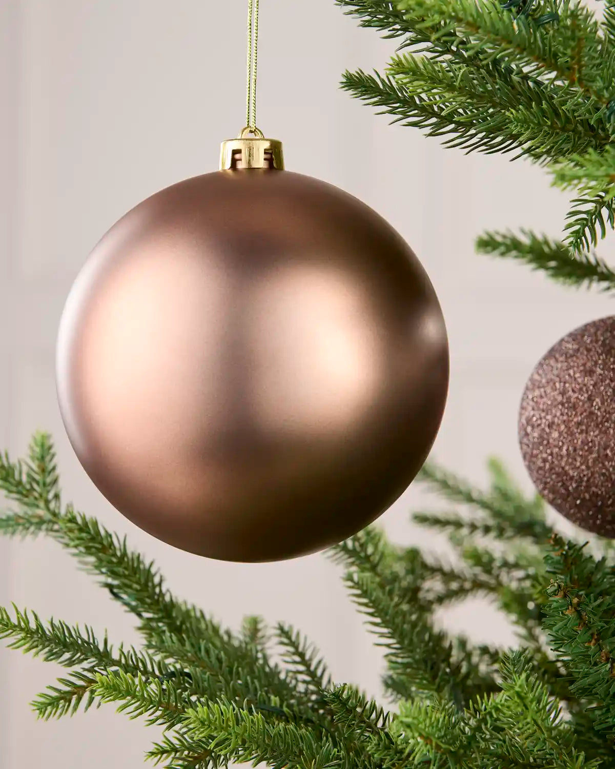 A large matte metallic Mocha Christmas bauble hanging from a tree branch, with green pine needles and part of a glittery ornament visible in the background.