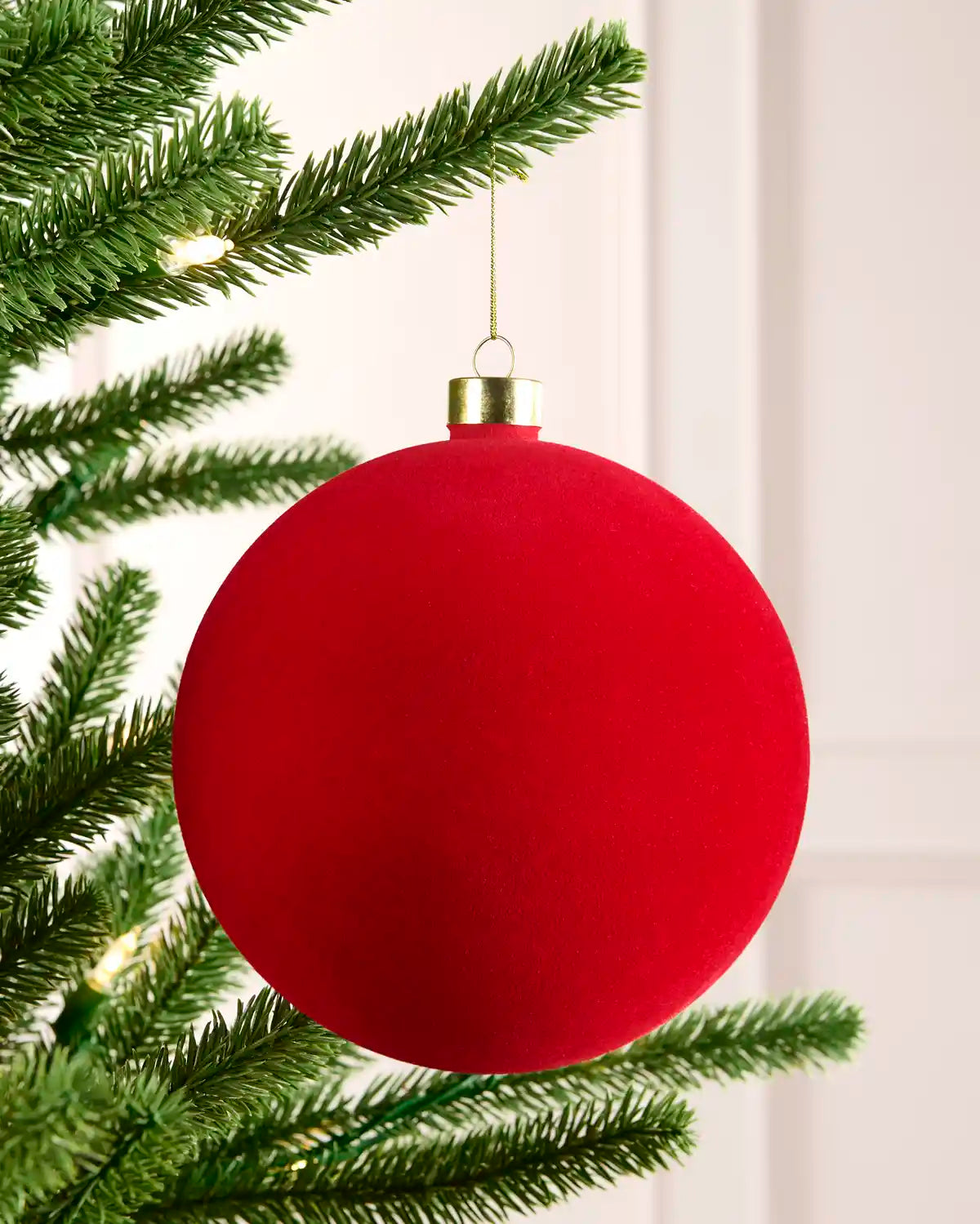 A large matte red Christmas tree ornament hanging from a green pine tree branch with warm white string lights in the background.