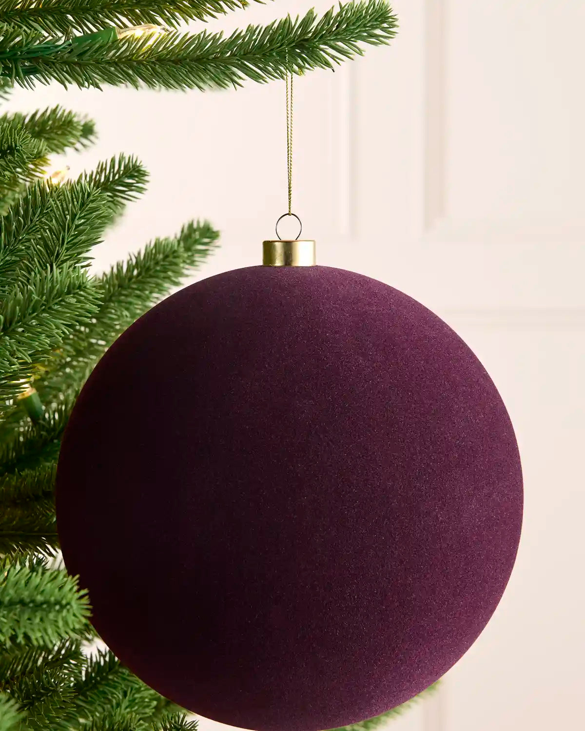 A large purple velvet Christmas tree ornament hanging from a green pine tree branch with a gold cap and gold hanging string.