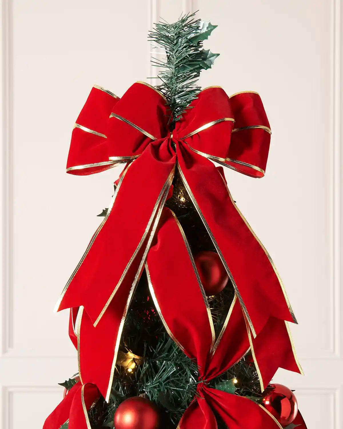 A large red Christmas tree bow with gold trim, tied to a decorated Christmas tree with red ornaments and lights, set against a light cream panneled wall.