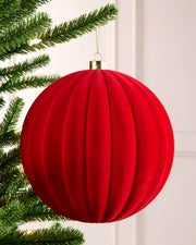 A large red ribbed velvet Christmas tree ornament hanging from a green pine tree branch with warm string lights in the background.
