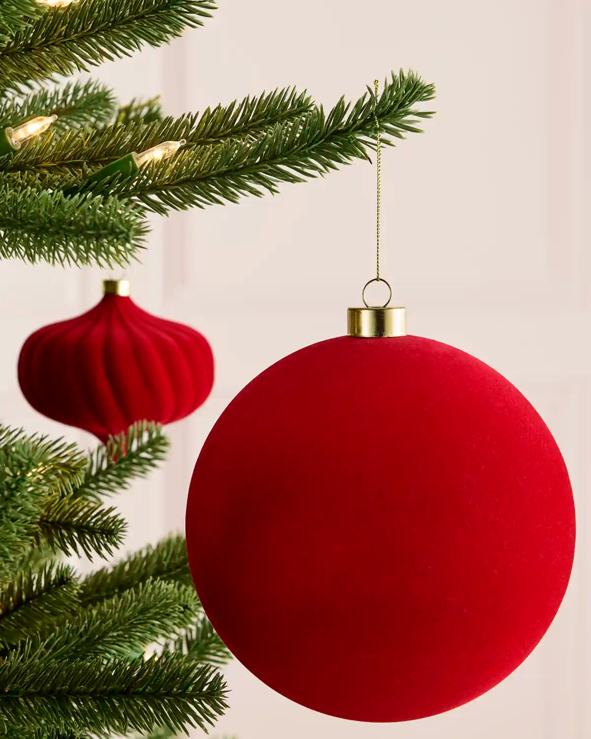 A large red spherical Christmas ornament with a gold cap hangs from a green Christmas tree branch, with a smaller matching red ornament blurred in the background.