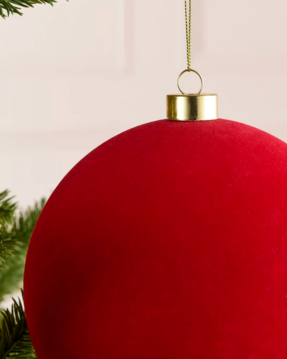 A large red velvet Christmas bauble ornament hanging from a gold cap and gold chain, with green pine branches visible at the edges.