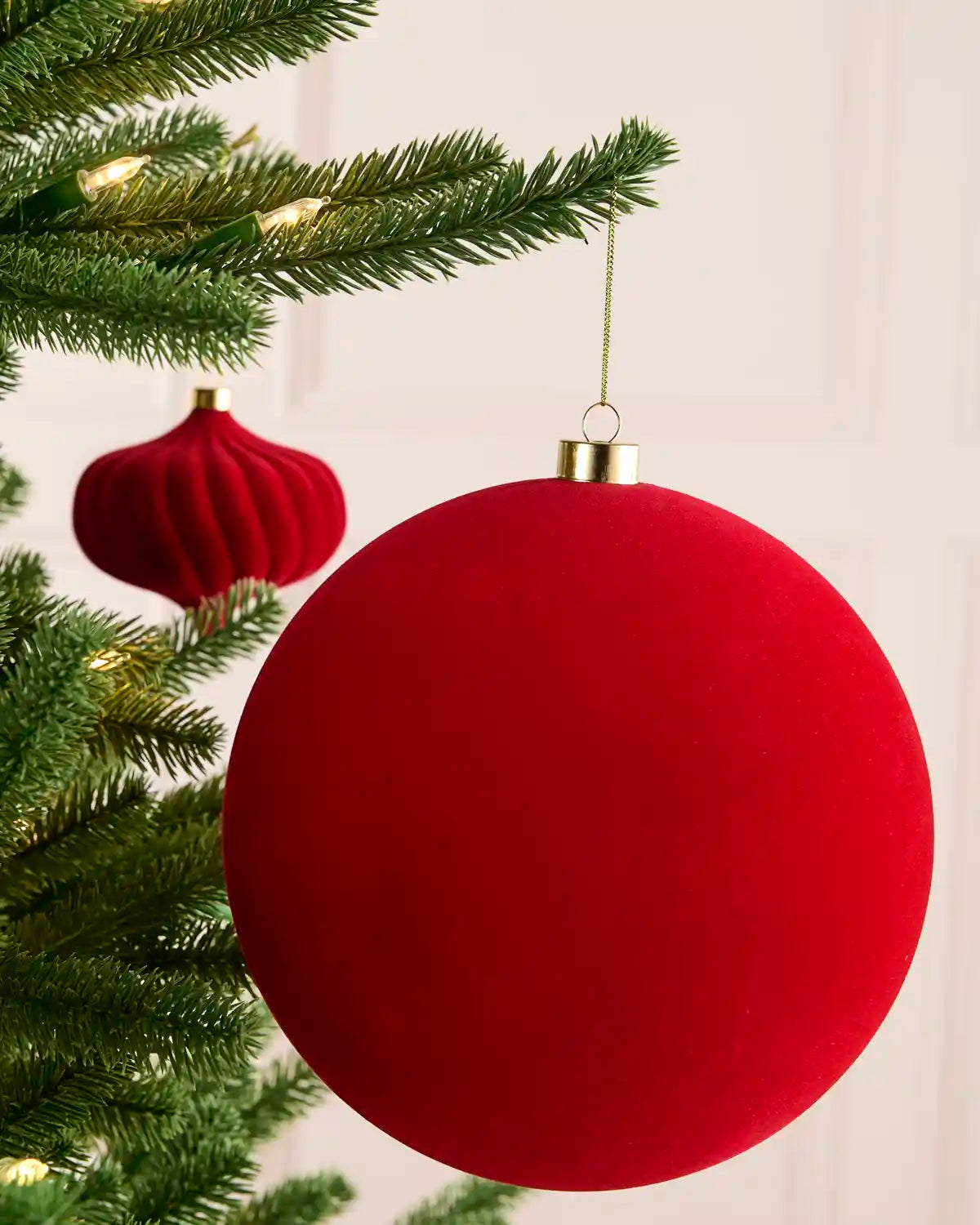 A large red velvet Christmas bauble ornament hanging from a green Christmas tree branch, with a smaller matching red ornament blurred in the background.
