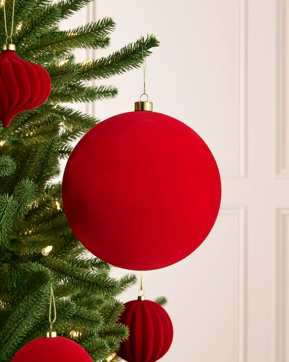 A large red velvet Christmas bauble ornament hanging from a green Christmas tree with warm white string lights, alongside other red paper lantern ornaments against a white paneled wall.