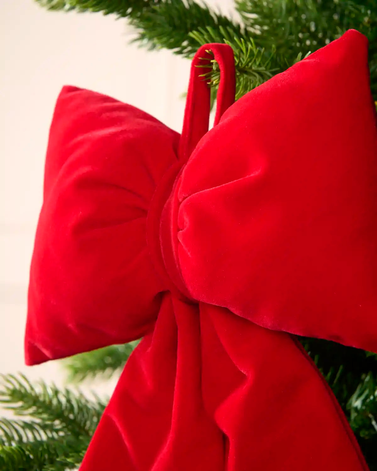 A large red velvet bow Christmas tree topper with a hanging loop, shown hanging from a green Christmas tree branch against a light background.