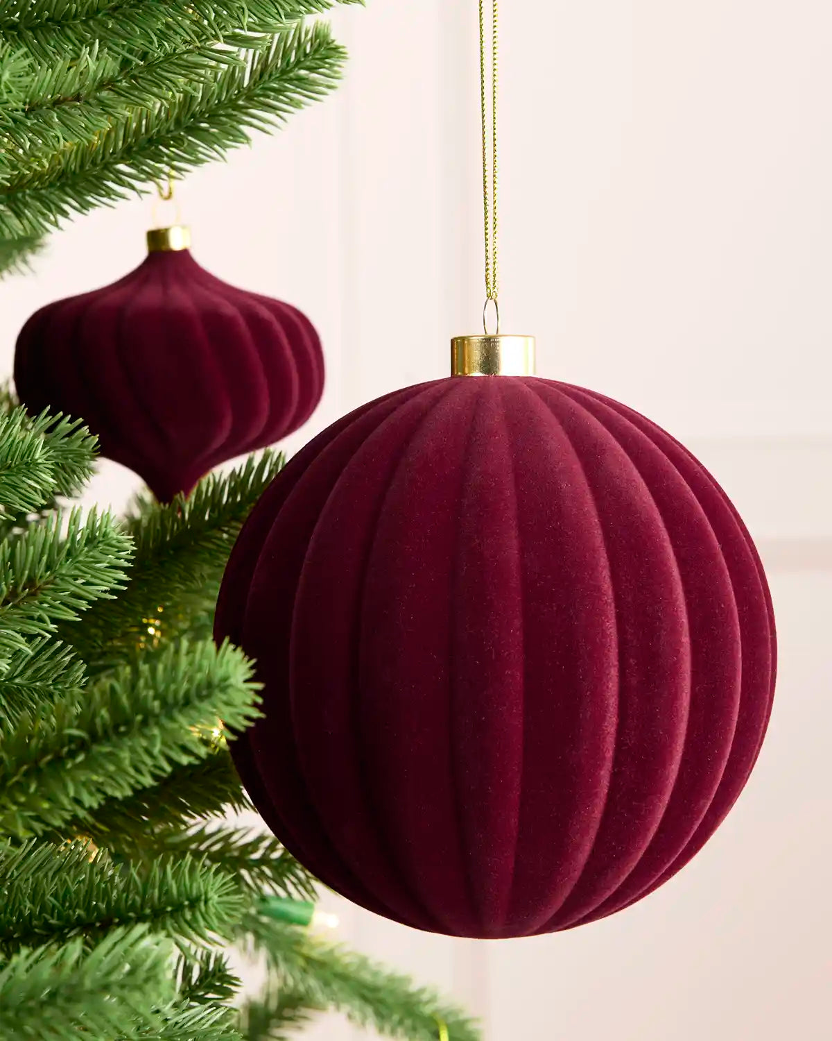 A large ribbed velvet Christmas ornament in deep burgundy hanging from a gold string next to green pine tree branches, with a second matching ornament blurred in the background.