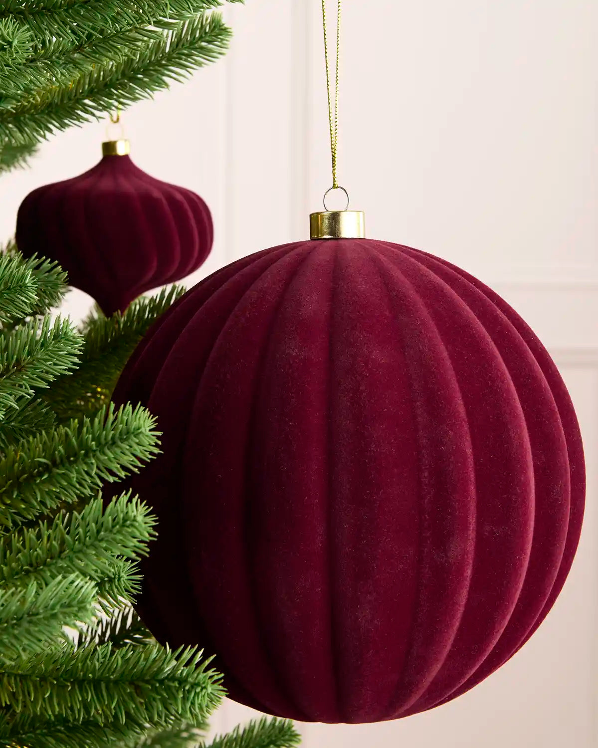 A large ribbed velvet Christmas tree ornament in burgundy hanging from a gold loop, shown next to green pine tree branches against a light neutral wall.