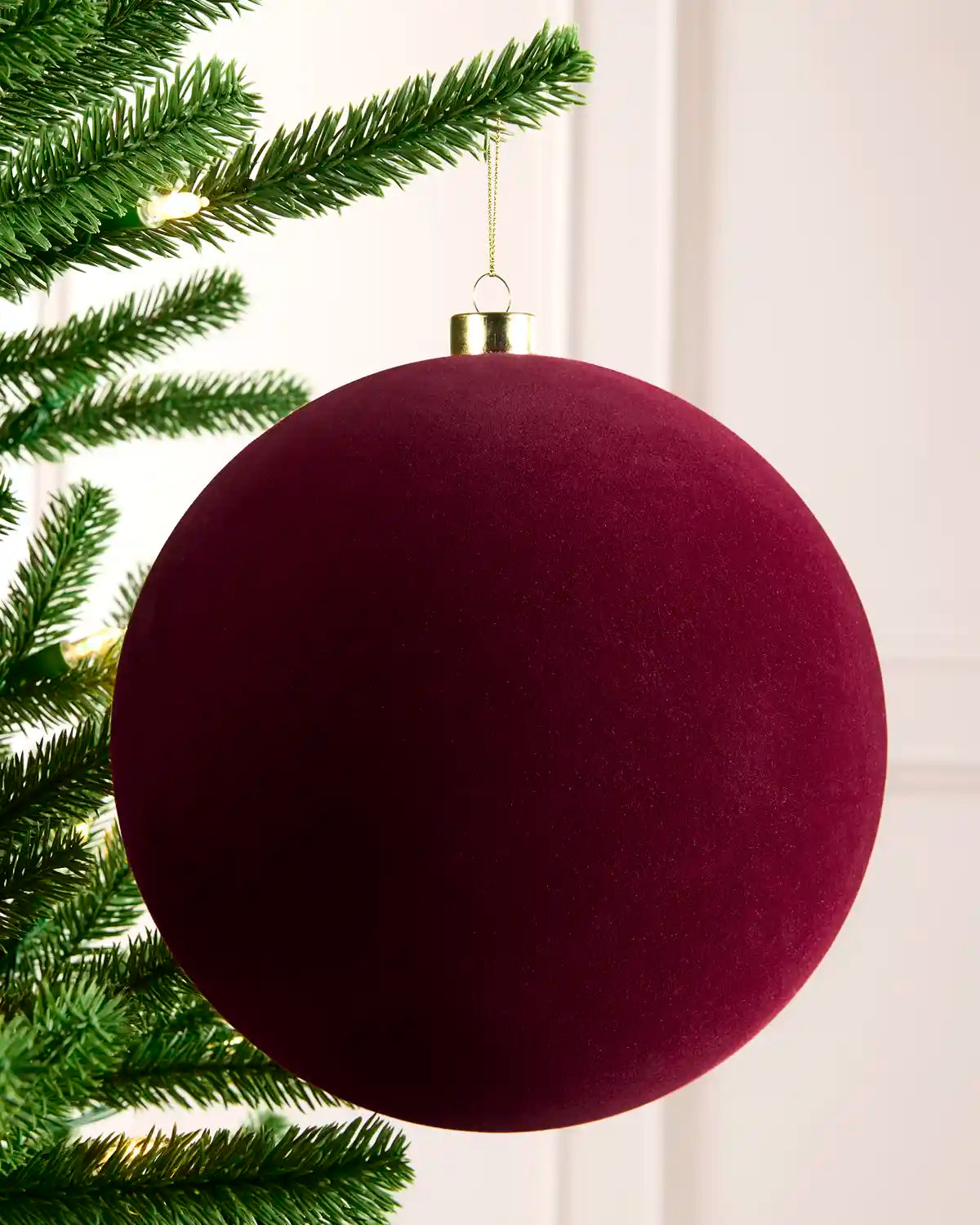 A large round Christmas ornament in deep burgundy velvet hanging from a green Christmas tree branch against a white paneled wall.