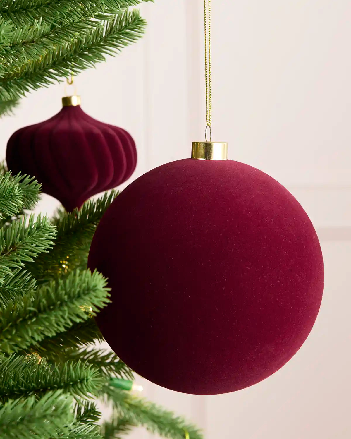 A large round burgundy velvet Christmas ornament hanging from a gold cap on a gold string, displayed next to green pine tree branches. A second matching ornament hangs in the background.