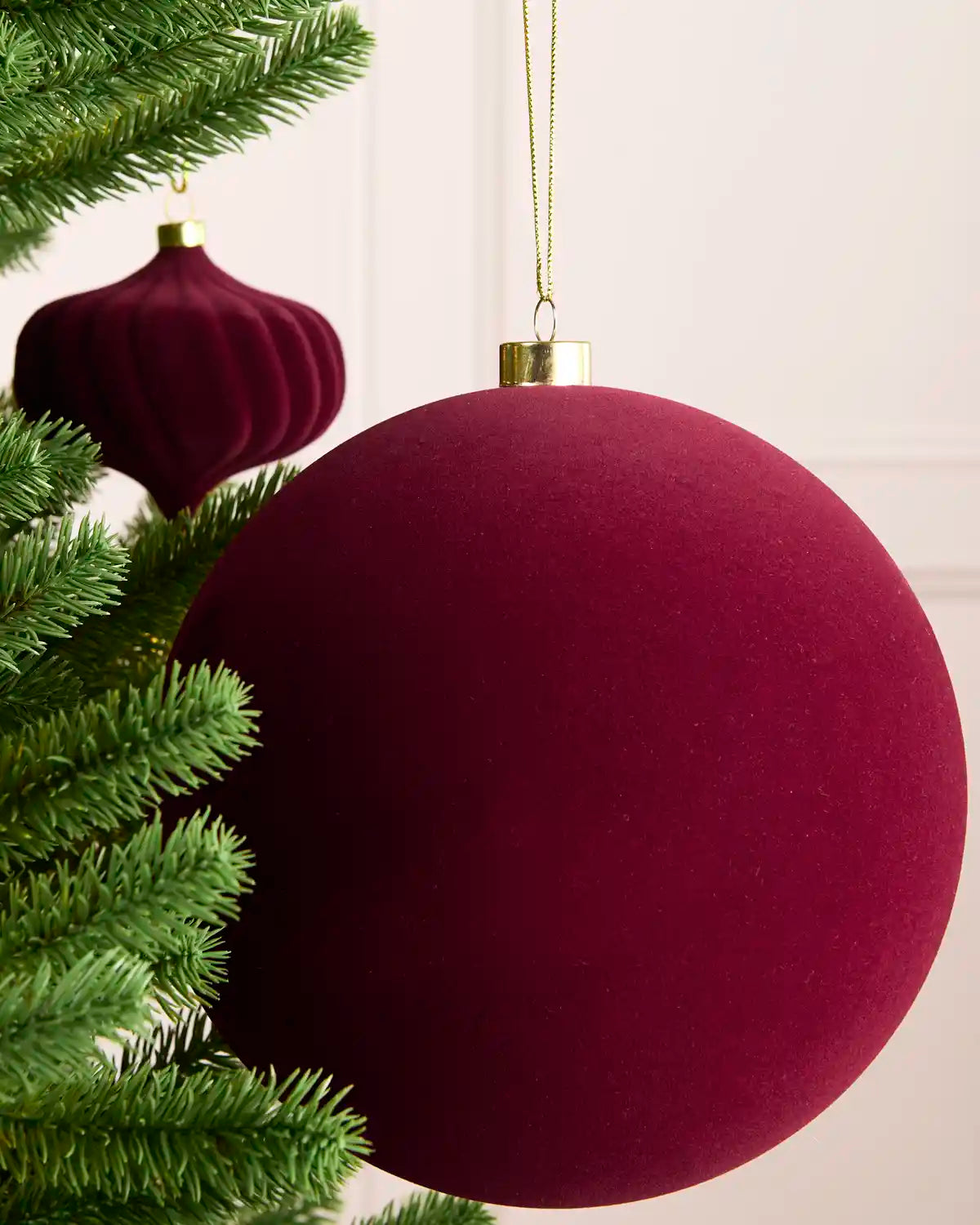 A large round burgundy velvet Christmas ornament hanging on a green Christmas tree, with a smaller matching ornament in the background.