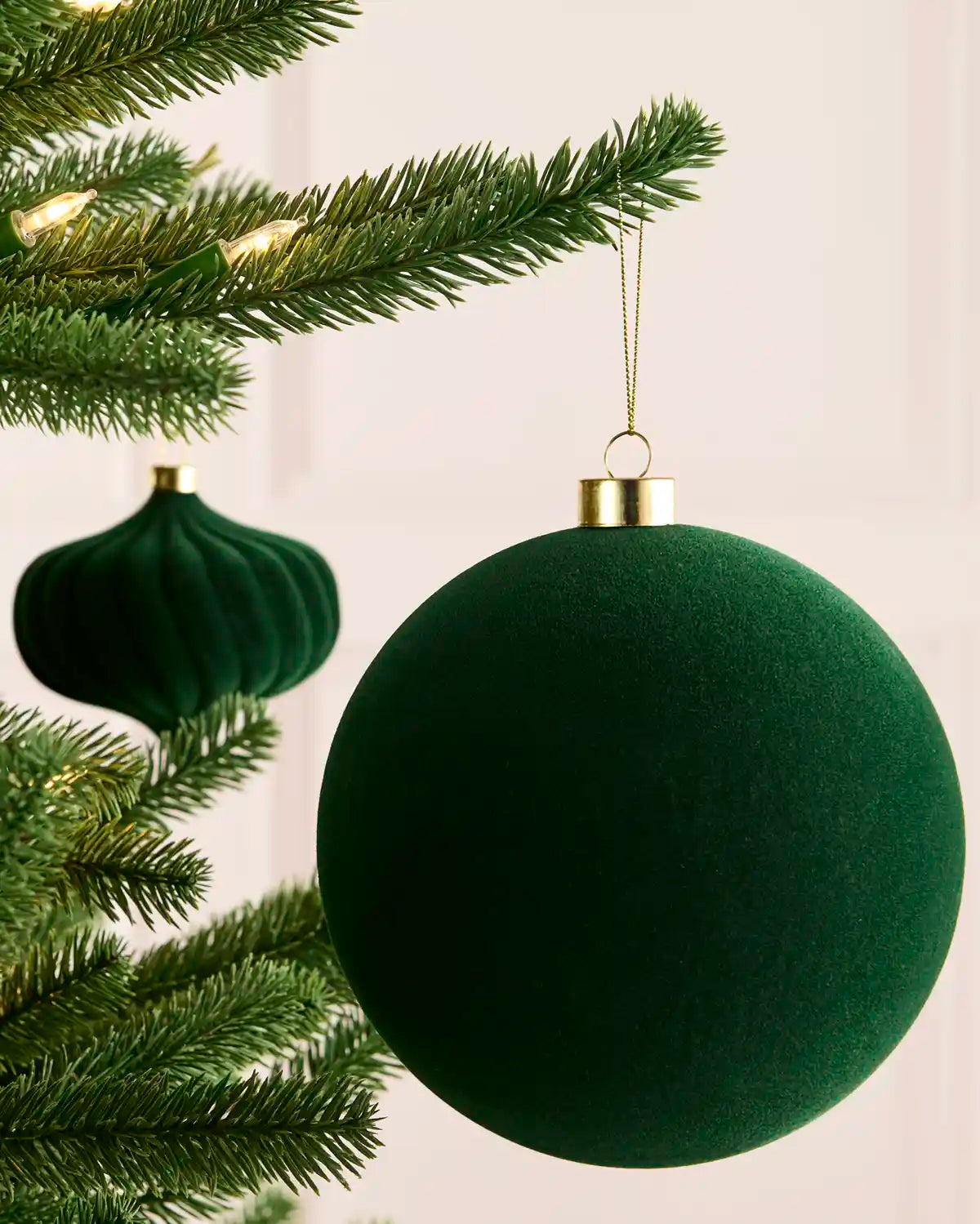 A sage green velvet Christmas bauble ornament hanging from a gold cap on a pine tree branch, with a second matching ornament blurred in the background.