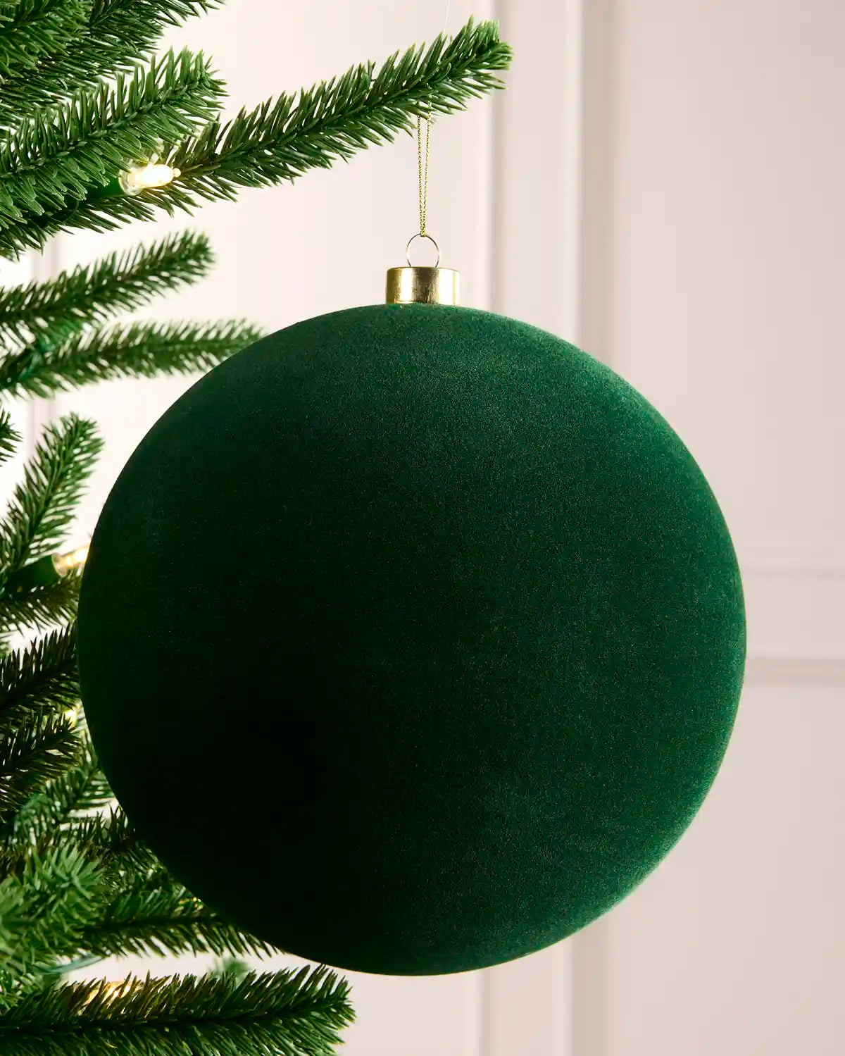 A large sage  green velvet Christmas tree ornament hanging from a green pine tree branch against a light off-white paneled wall.