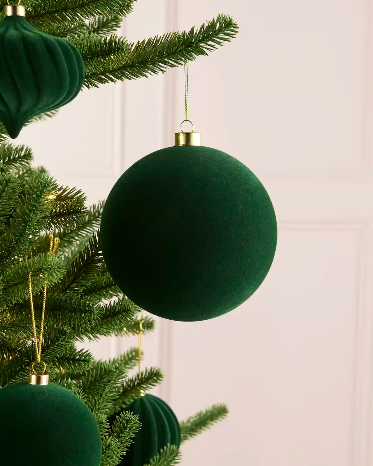 A large sage green velvet Christmas tree ornament hanging from a green pine tree branch against a pale panelled wall.