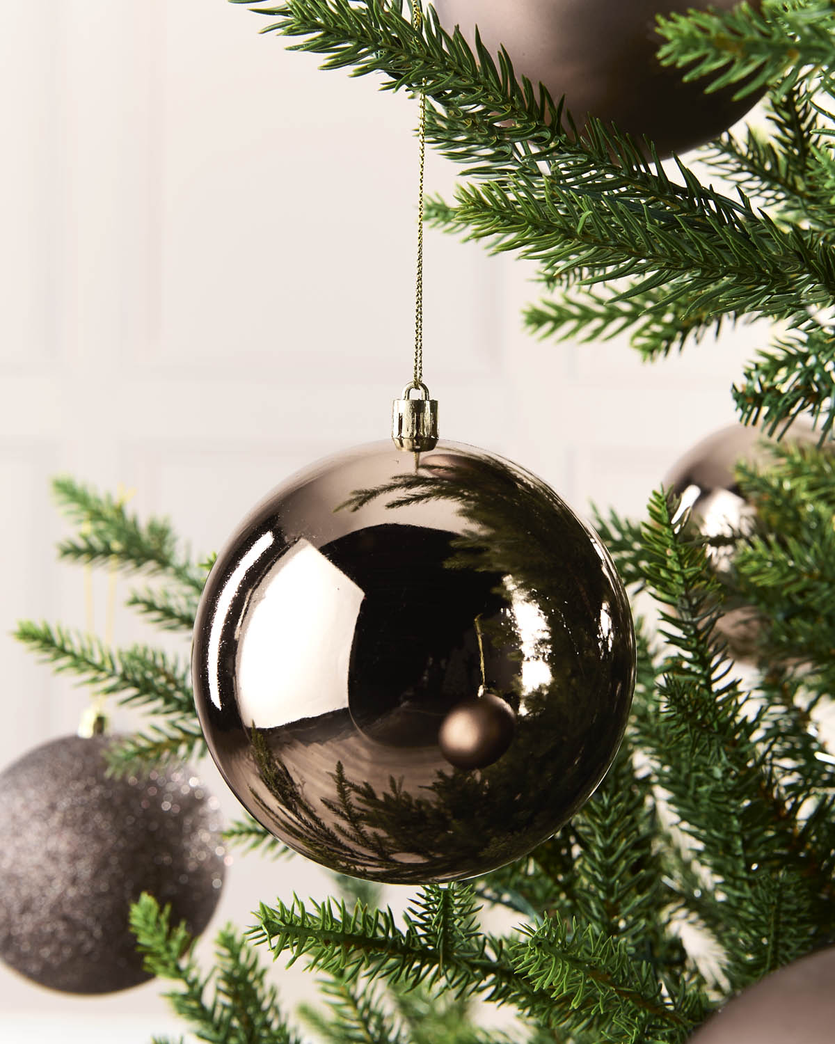 A large, shiny metallic Mocha Christmas ball ornament hanging from a green pine tree branch, with other ornaments and a softly blurred background visible.