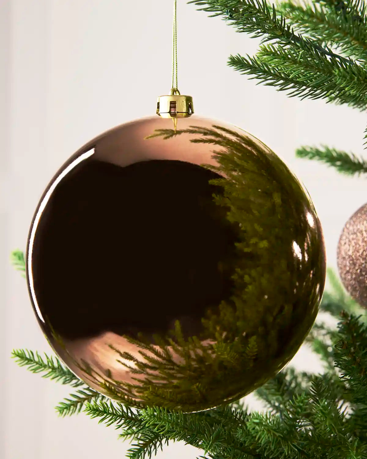 A large, shiny metallic Mocha Christmas bauble hanging from a green pine tree branch against a soft white background.