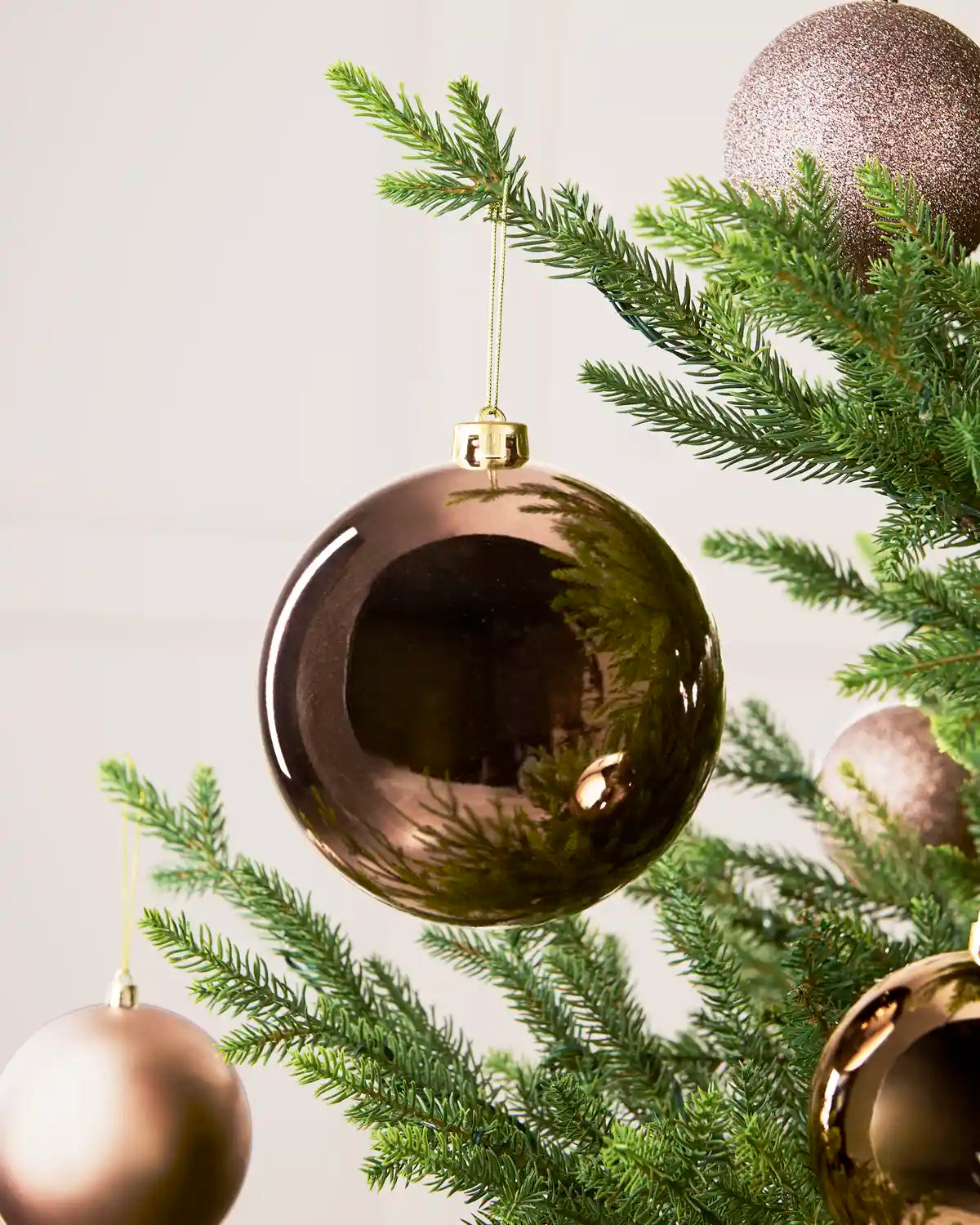 A large, shiny metallic Mocha Christmas bauble ornament hanging from a green pine tree branch, with other matching bronze ornaments and pine needles in the background.