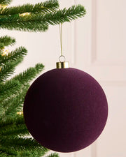 A large, traditional round purple Christmas tree ornament hanging from a green pine tree branch with warm string lights in the background.