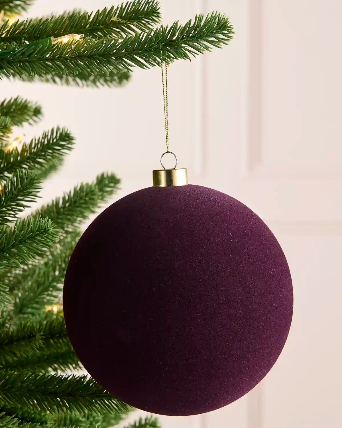 A large, traditional round purple Christmas tree ornament hanging from a green pine tree branch with warm string lights in the background.