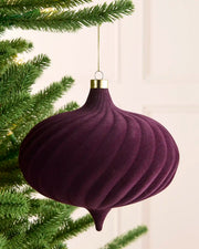 A large velvet purple spiral bauble hanging on a Christmas tree with gold string against a light background