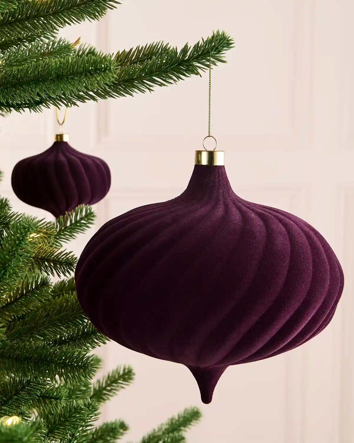 A large velvet purple spiral bauble hanging on a Christmas tree with gold string against a light background and a smaller matching bauble 