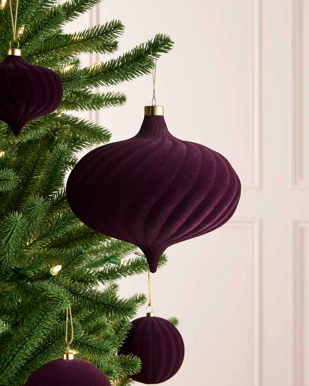 A large velvet purple spiral bauble hanging on a Christmas tree with gold string against a light background and smaller matching baubles  