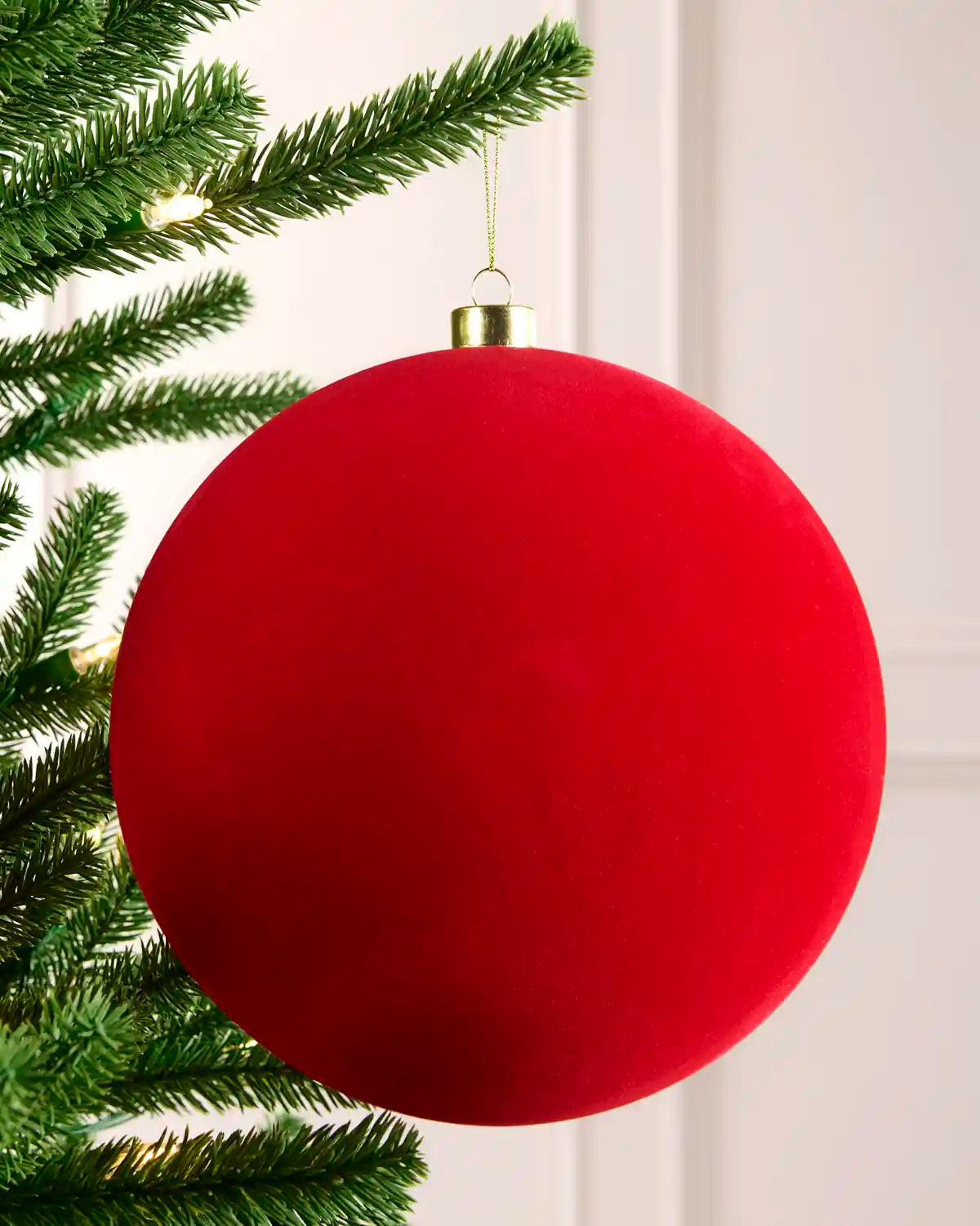 A large, velvet red spherical Christmas ornament hanging from a green Christmas tree branch against a white panelled wall.