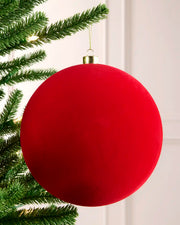 A large, velvet red spherical Christmas ornament hanging from a green Christmas tree branch against a white panelled wall.