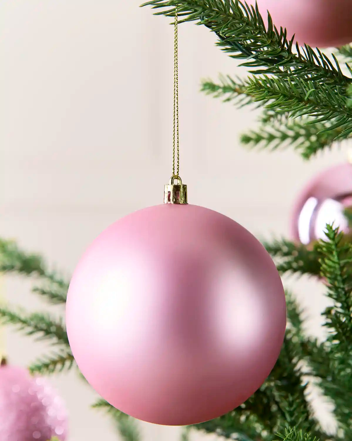 A matt pink Christmas bauble bauble hanging from a gold string on a green Christmas tree branch.