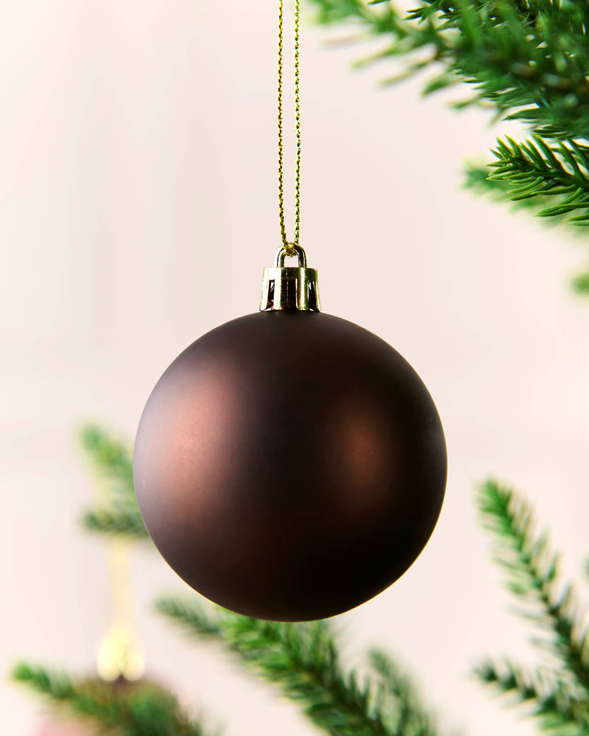 A matte Espresso Christmas bauble ornament hanging from a gold string against a soft light background with blurred green pine branches.