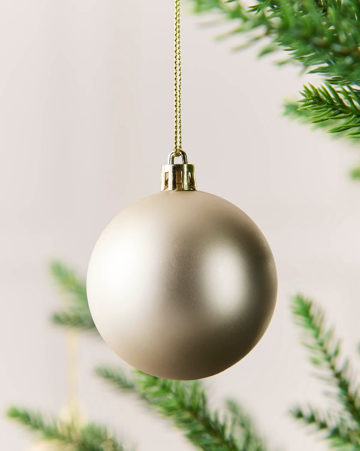 A matte Latte Christmas bauble hanging from a gold string against a soft white background with blurred green pine branches.