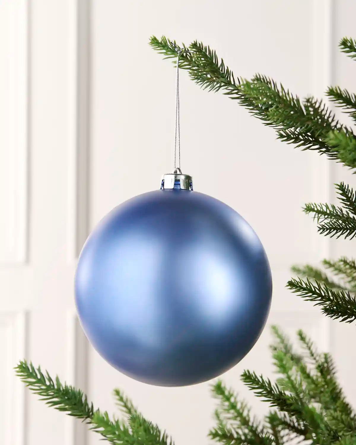 A matte blue Christmas tree ornament hanging from a silver cap by a thin string, framed by green pine branches against a soft off-white paneled background.