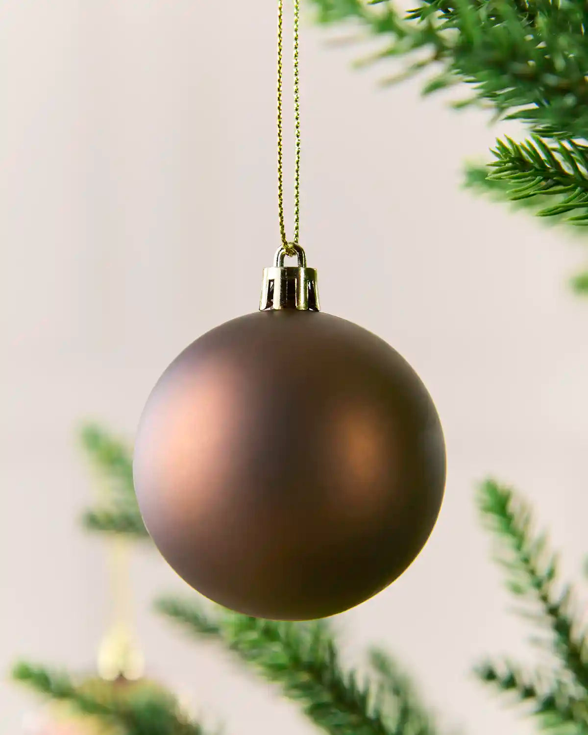 A matte dark Mocha Christmas ornament hanging from a gold string against a soft light background with blurred green pine branches.