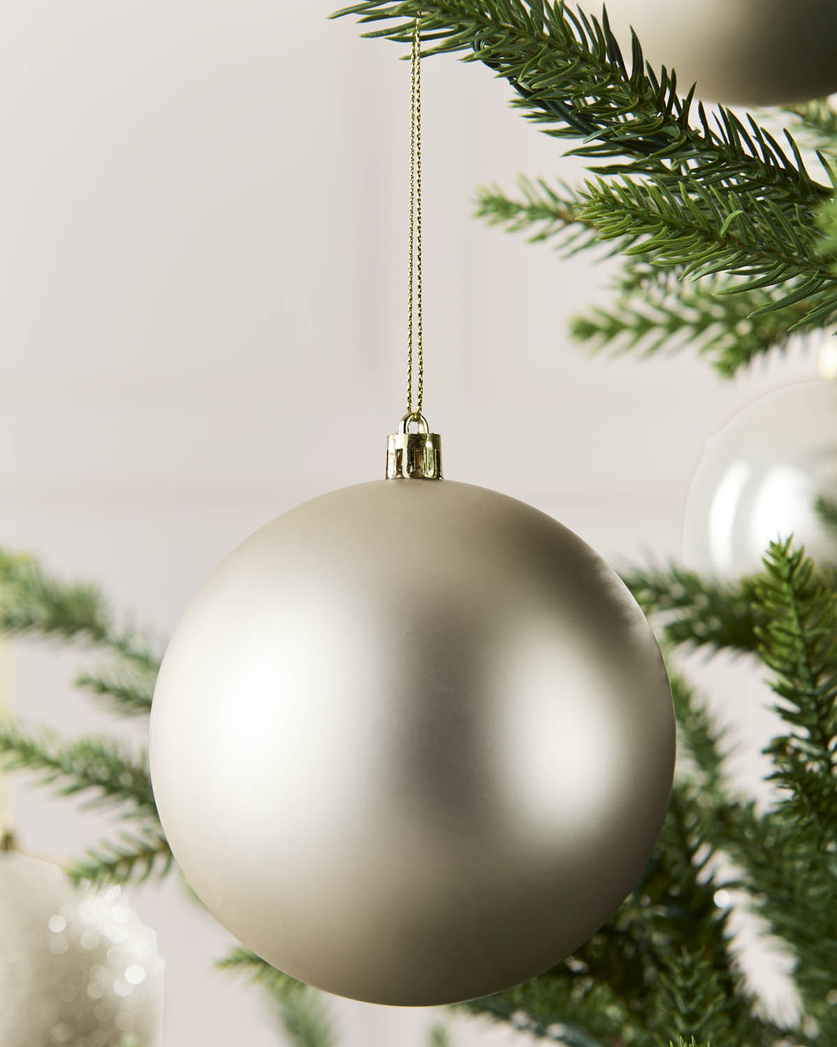 A matte latte Christmas bauble hanging from a gold string against a soft neutral background, with green pine branches and other ornaments partially visible.