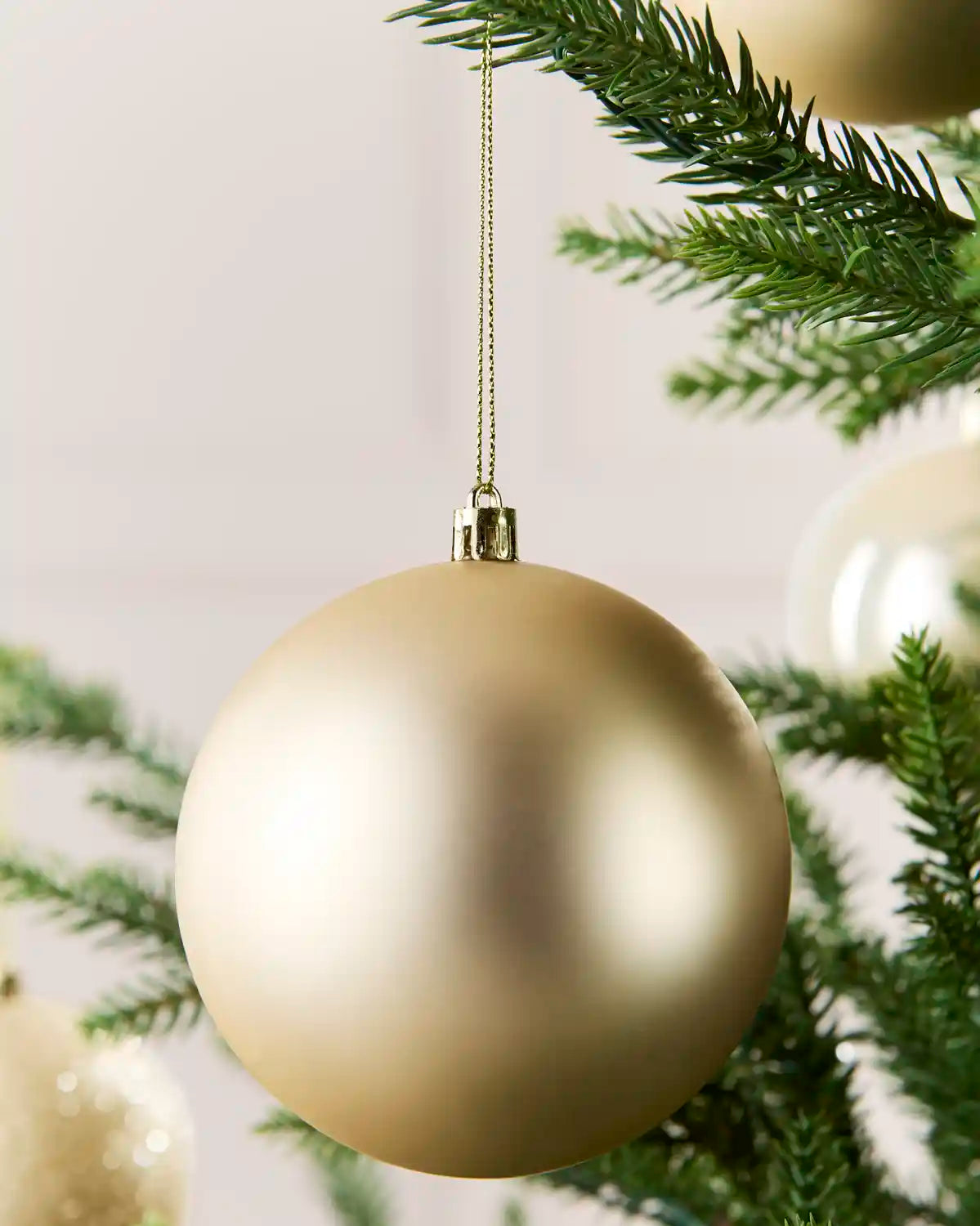 A matte latte Christmas bauble hanging from a gold string against a soft neutral background, with green pine branches and other ornaments partially visible.
