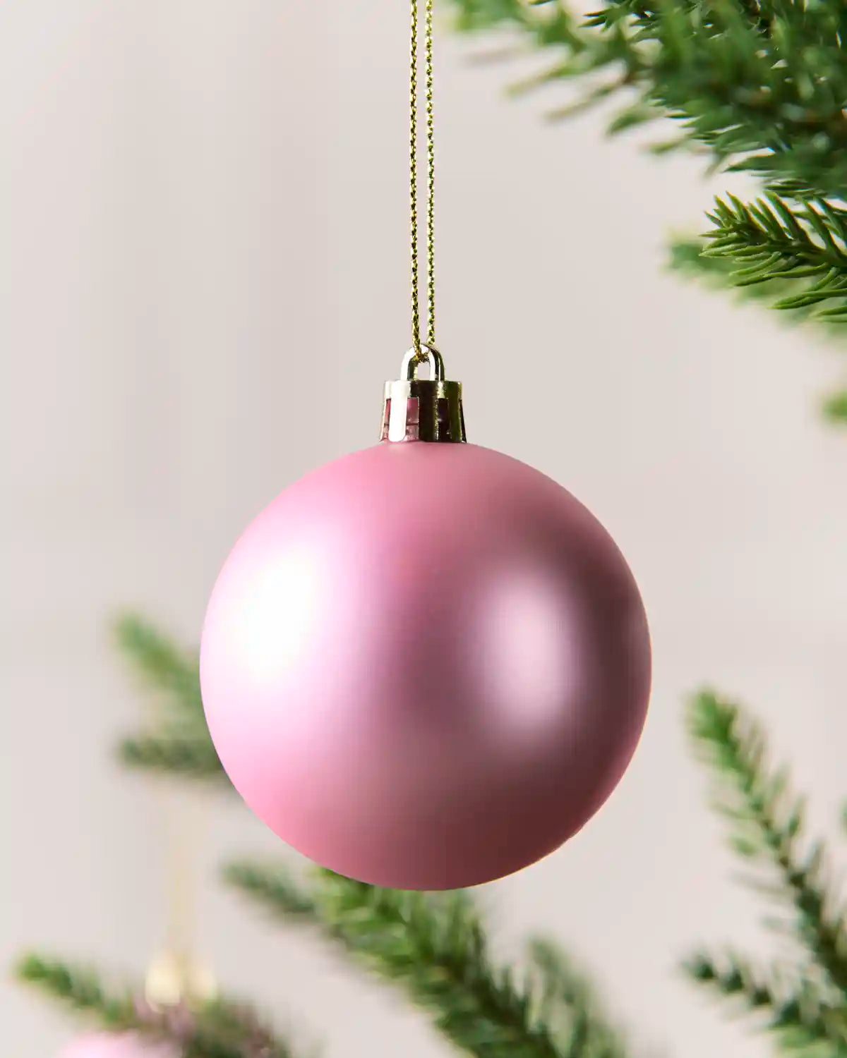 A matte light pink Christmas bauble ornament hanging from a gold-colored chain against a soft, blurred background with green pine branches.