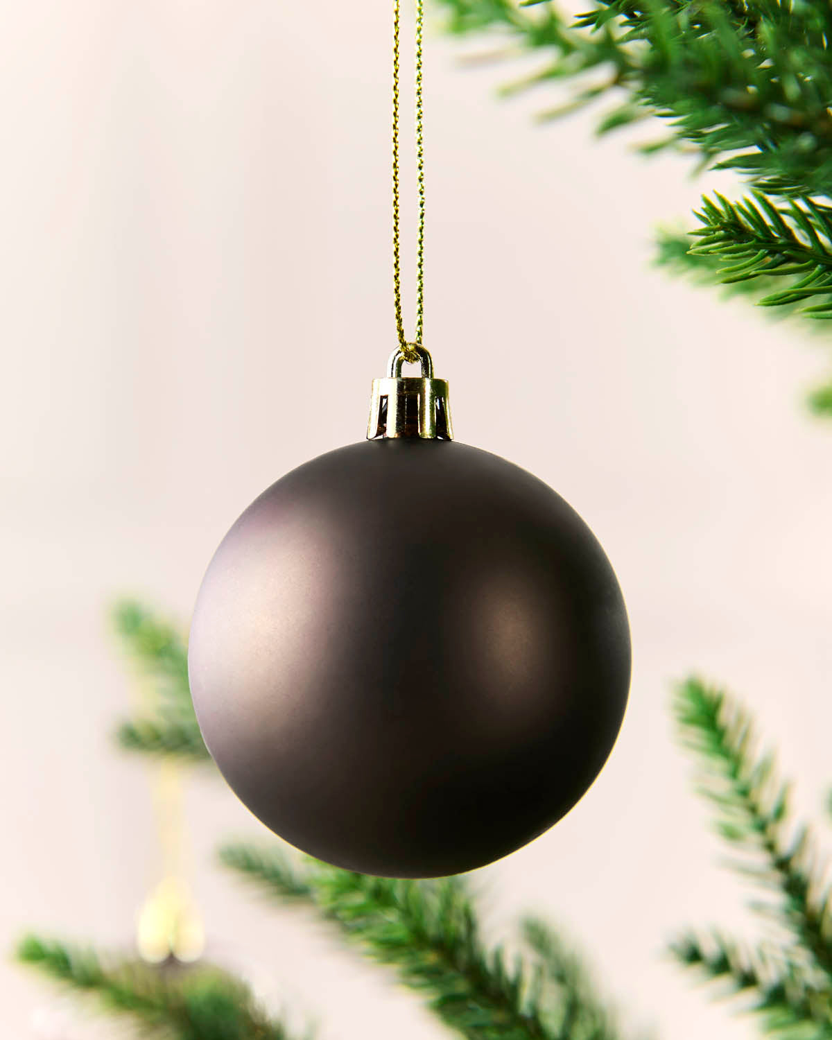A matte mocha Christmas ball ornament hanging from a gold string, with blurred green pine branches in the background.
